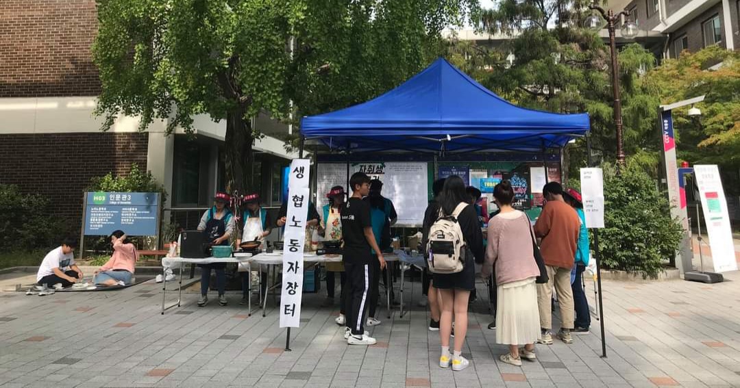 2019년 가을 생협 노동자 파업 과정에서 찍힌 생협 노동자 장터 사진이다. 파업 당시 노동자들이 인문관3 건물 앞에 “생협 노동자 장터”라는 팻말을 내건 천막을 치고 연대장터를 운영하며 학생들에게 음식을 판매하는 모습이다. 장터 천막 앞에 음식을 구매하려는 학생들이 줄을 서 있고, 옆에는 돗자리를 깔고 취식하는 학생들의 모습이 보인다.