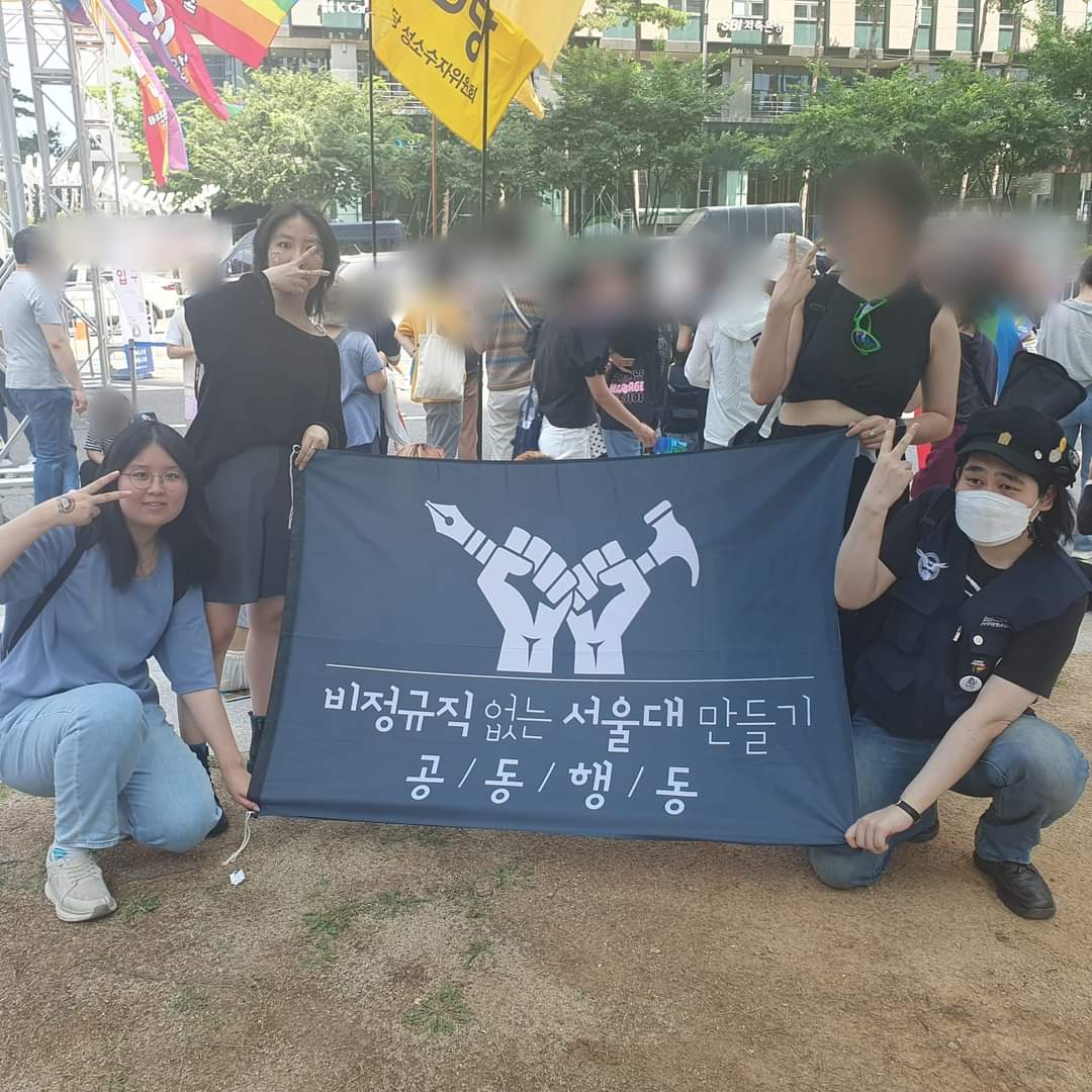 비서공 회원들이 검정색 비서공 깃발의 네 귀퉁이를 잡고 V자 포즈를 취하고 있다. 비서공 깃발에는 두 개의 주먹이 각각 펜과 망치를 들고 있다.