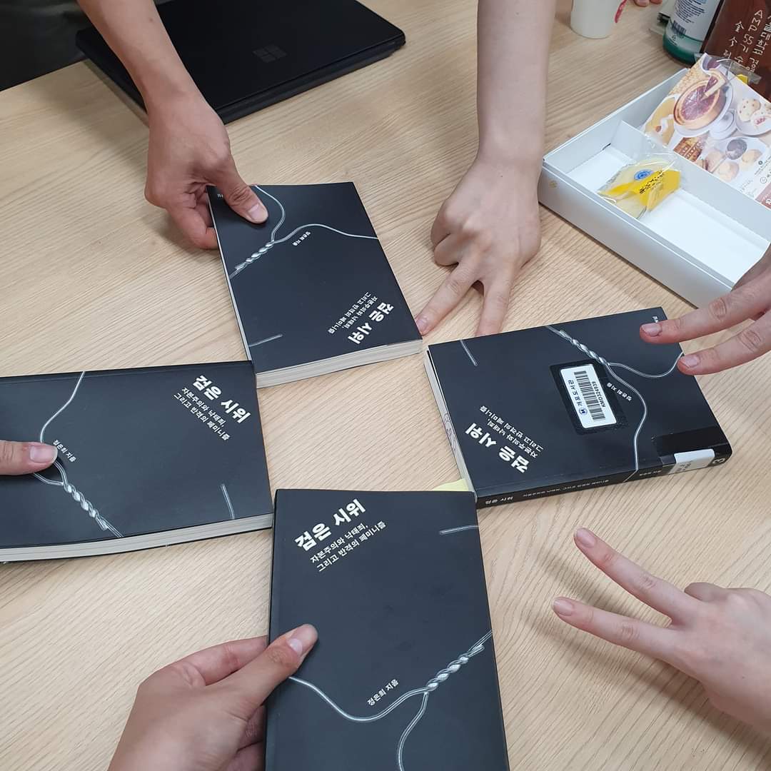 옅은 갈색의 책상 위에 4권의 책이 돌아가며 놓여 있고, 6명의 손이 책을 잡거나 혹은 브이 자로 포즈를 취하고 있는 모습을 위에서 찍은 사진이다. 4권의 책은 검은 표지에 흰색으로 포궁의 모습을 형상화한 도안이 위치하고 있고, “검은 시위 - 자본주의와 낙태죄, 그리고 반격의 페미니즘 / 정은희 지음”이라고 흰 색의 글씨가 쓰인 모습이다. 책상의 상단에는 노트북과 다과 등이 위치하고 있다.