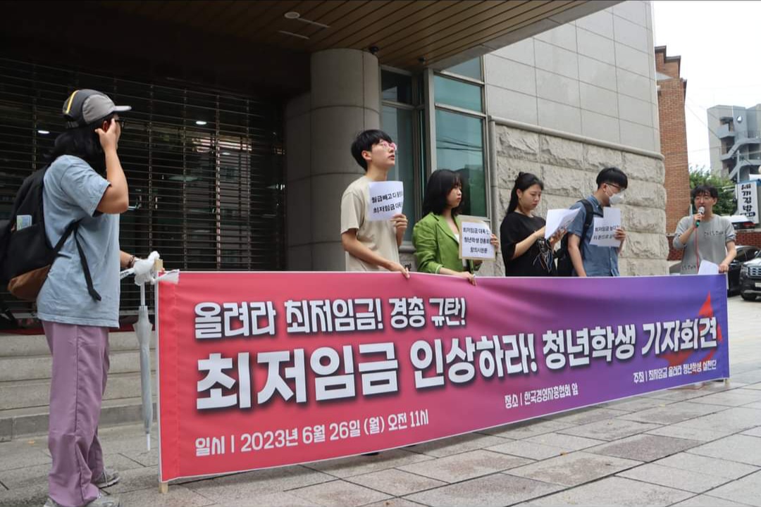 한 무리의 사람들이 현수막을 세워놓고 마이크를 들고 기자회견을 진행하고 있다. 현수막에는 “올려라 최저임금! 경총 규탄! / 최저임금 인상하라! 청년학생 기자회견 / 일시 | 2023년 6월 26일 (월) 오전 11시 / 장소 | 한국경영자총협회 앞 / 주최 | 최저임금 올려라 청년학생 실천단” 이라고 쓰여 있다.